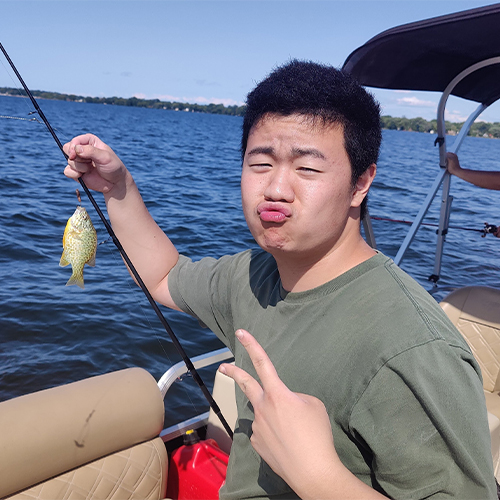 Denny on a pontoon boat, pulling a face and holding up a tiny sunfish he caught